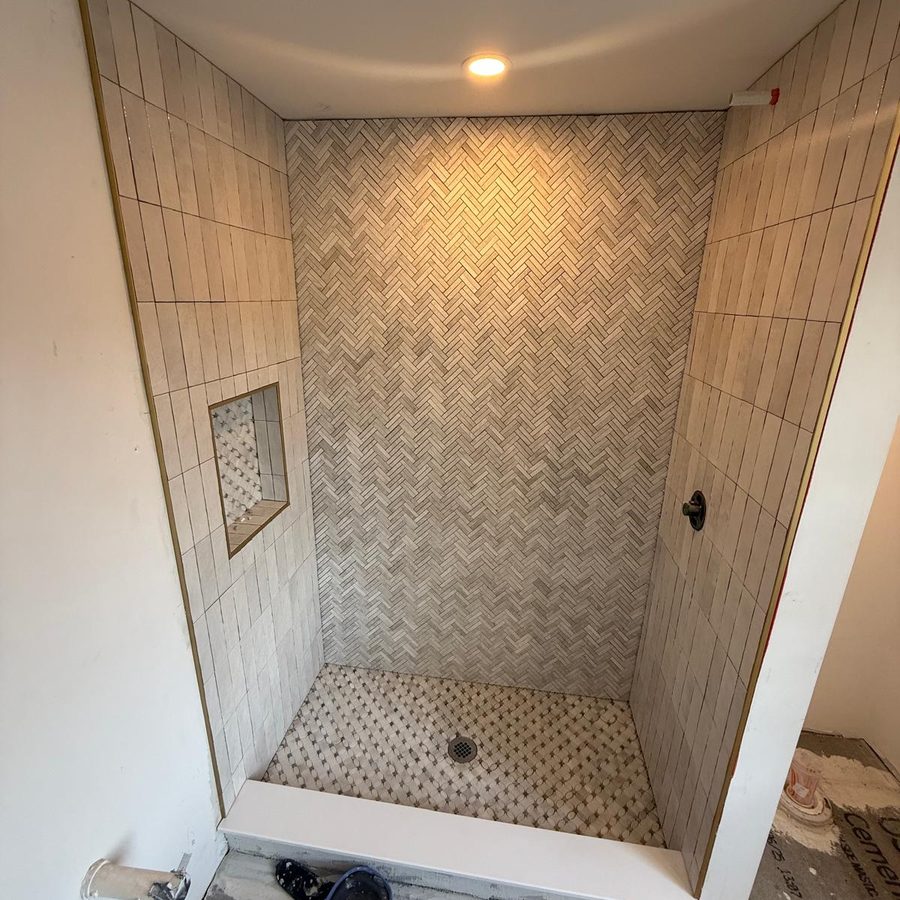 Walk-in shower with a herringbone back wall, vertical stacked tile, brass-trimmed niche and marble mosaic floor