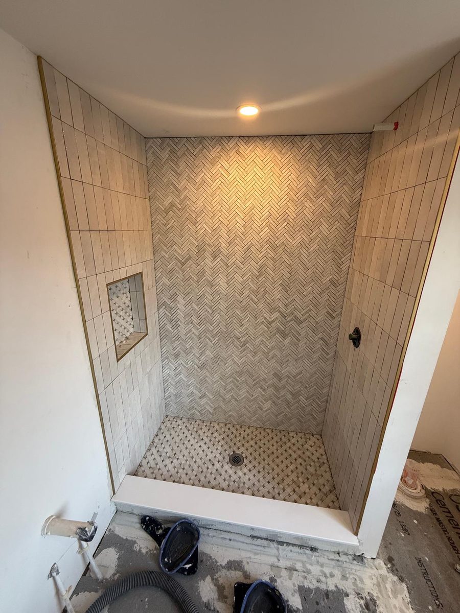 Walk-in shower with a herringbone back wall, vertical stacked tile, brass-trimmed niche and marble mosaic floor