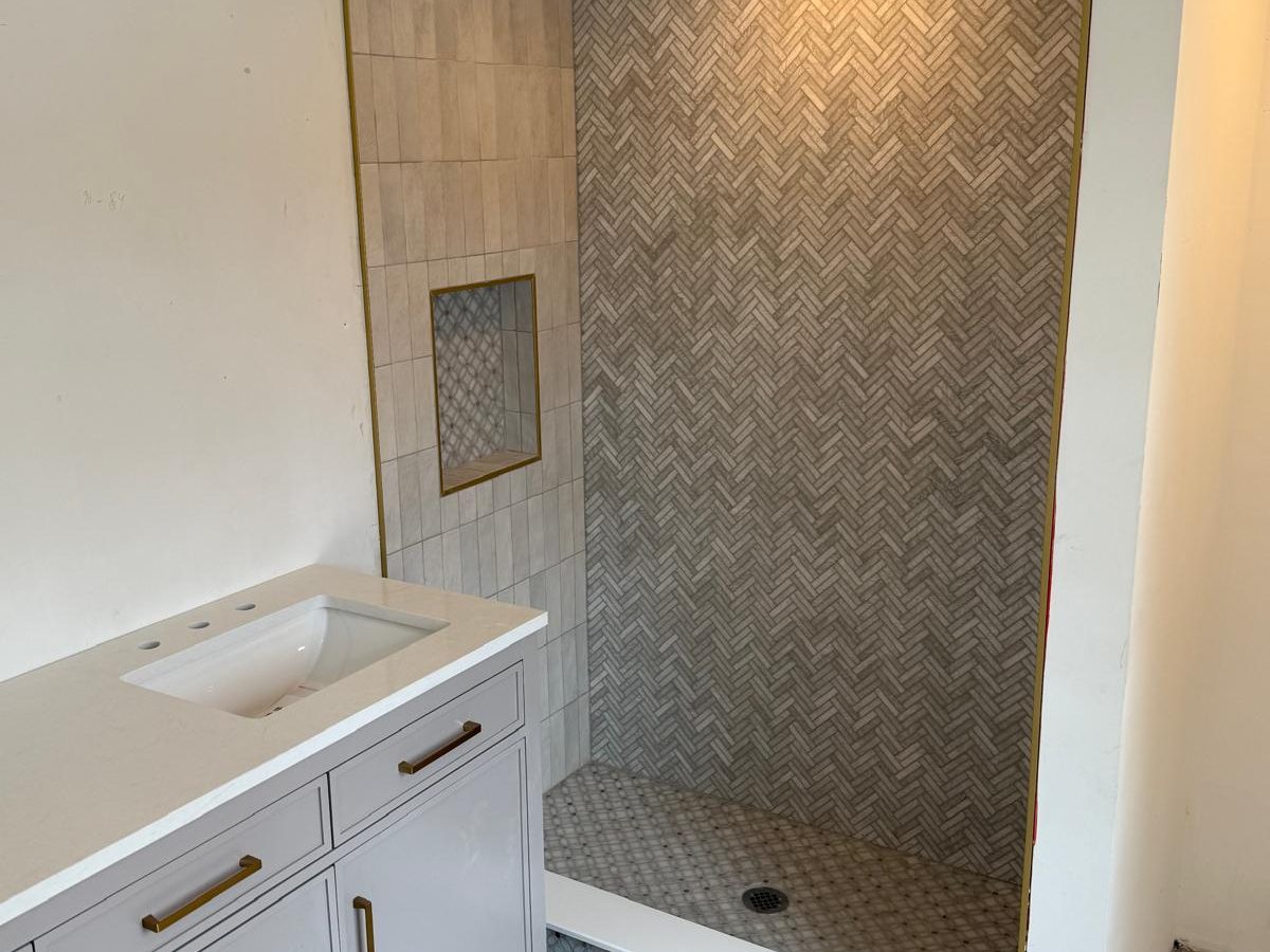 Finished bathroom with herringbone shower wall, brass-trimmed niche and a gray double vanity with brass pulls