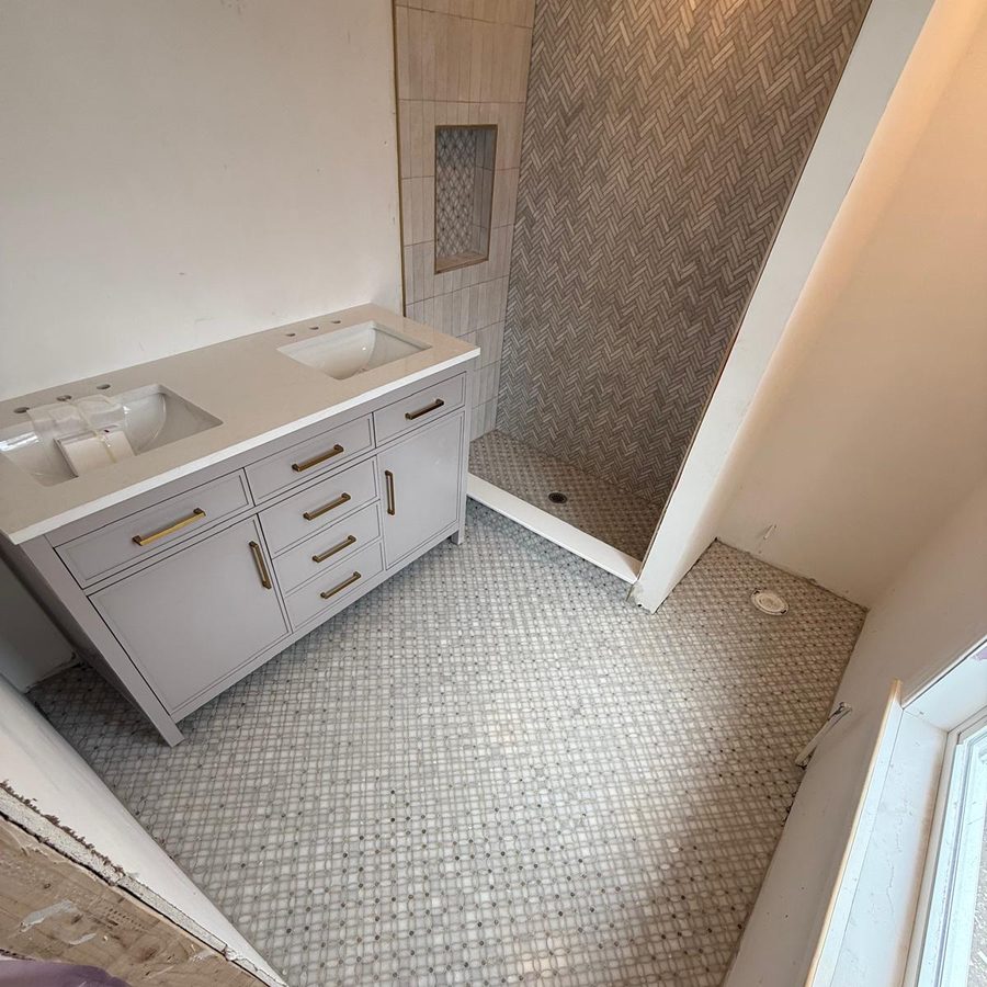 Gray double vanity with brass hardware on a marble mosaic floor beside the herringbone shower