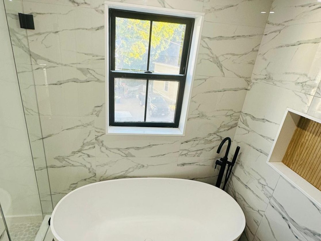 Freestanding tub under a black-framed window, with marble-look porcelain walls and a pebble mosaic floor