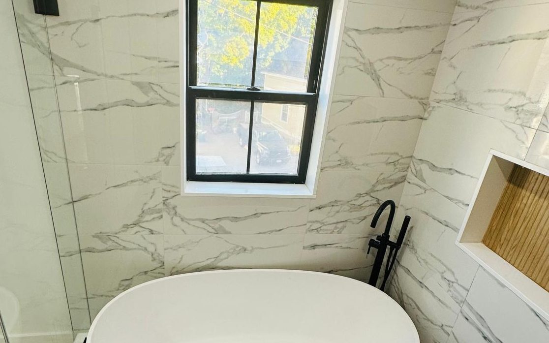 Freestanding tub under a black-framed window, with marble-look porcelain walls and a pebble mosaic floor