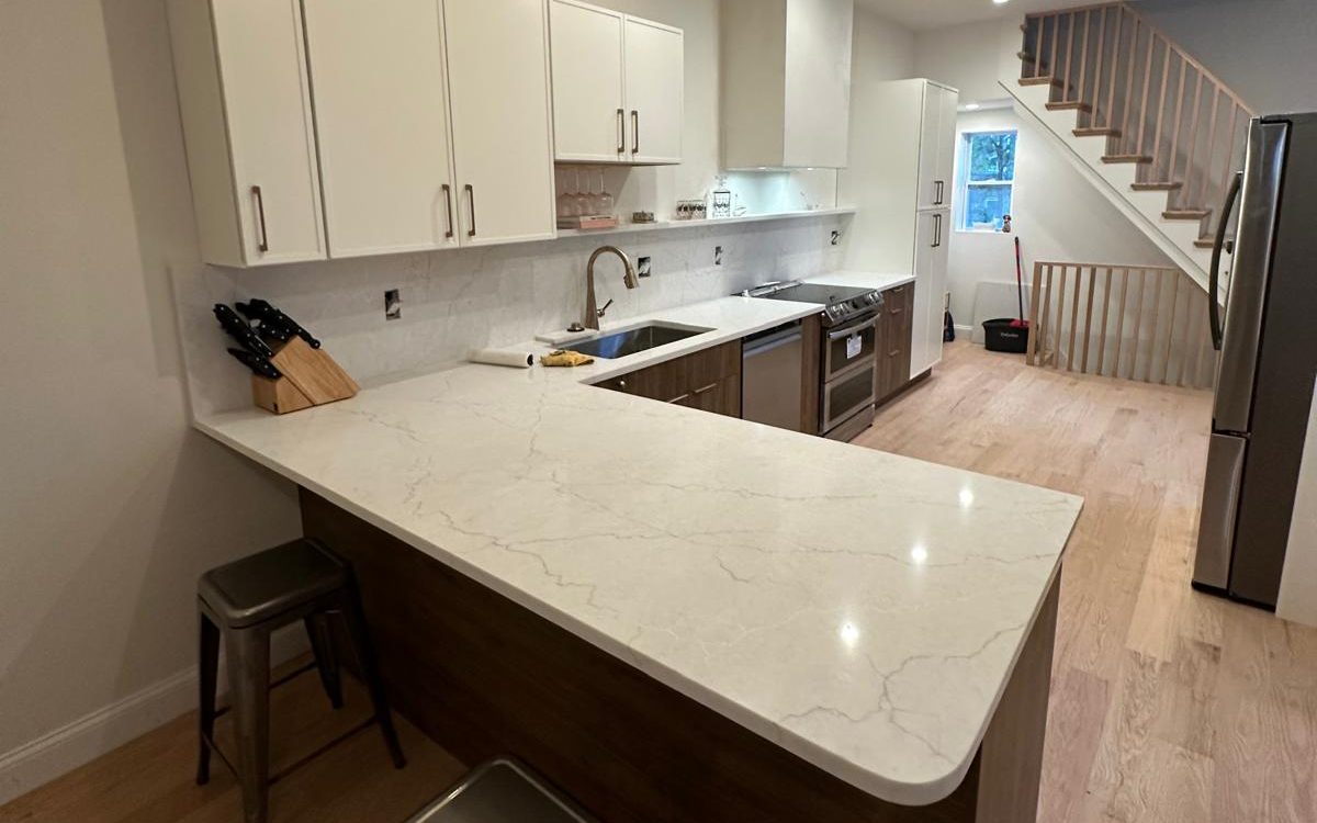 Townhouse kitchen with a quartz peninsula, white upper cabinets, walnut base cabinets and an oak stair beyond